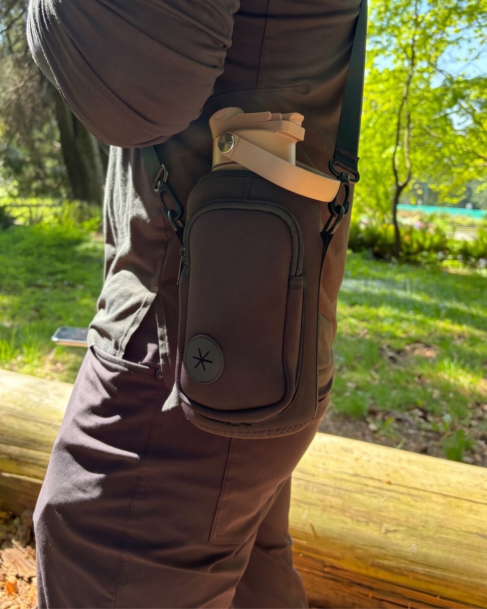 Close-up lifestyle shot of a person wearing the black OMNISE crossbody water bottle sling in a park, carrying a tan RoamWell bottle.