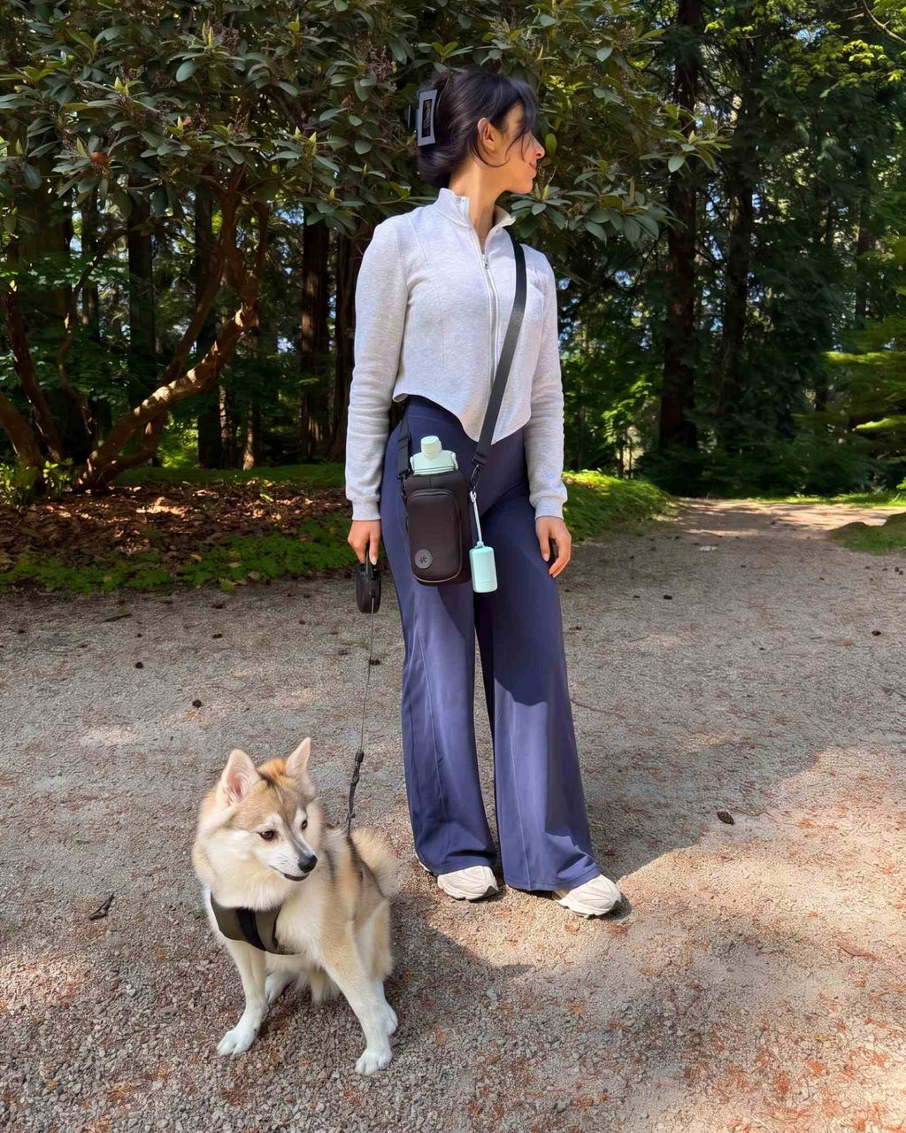 Girl walking her Pomsky on a forest trail while wearing the black OMNISE water bottle sling holding a light green RoamWell 32 bottle.
