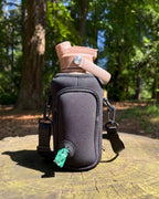 Black OMNISE water bottle sling resting on a tree stump in a forest, holding a tan, tidal sand RoamWell bottle. A green dog waste bag is being pulled through the built-in dispenser port on the front.