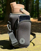 Black OMNISE water bottle sling resting on a forest tree stump, carrying a tan, tidal sand RoamWell bottle. The front zipper pocket is open, demonstrating its capacity to hold large smartphones like an iPhone 17 Pro.