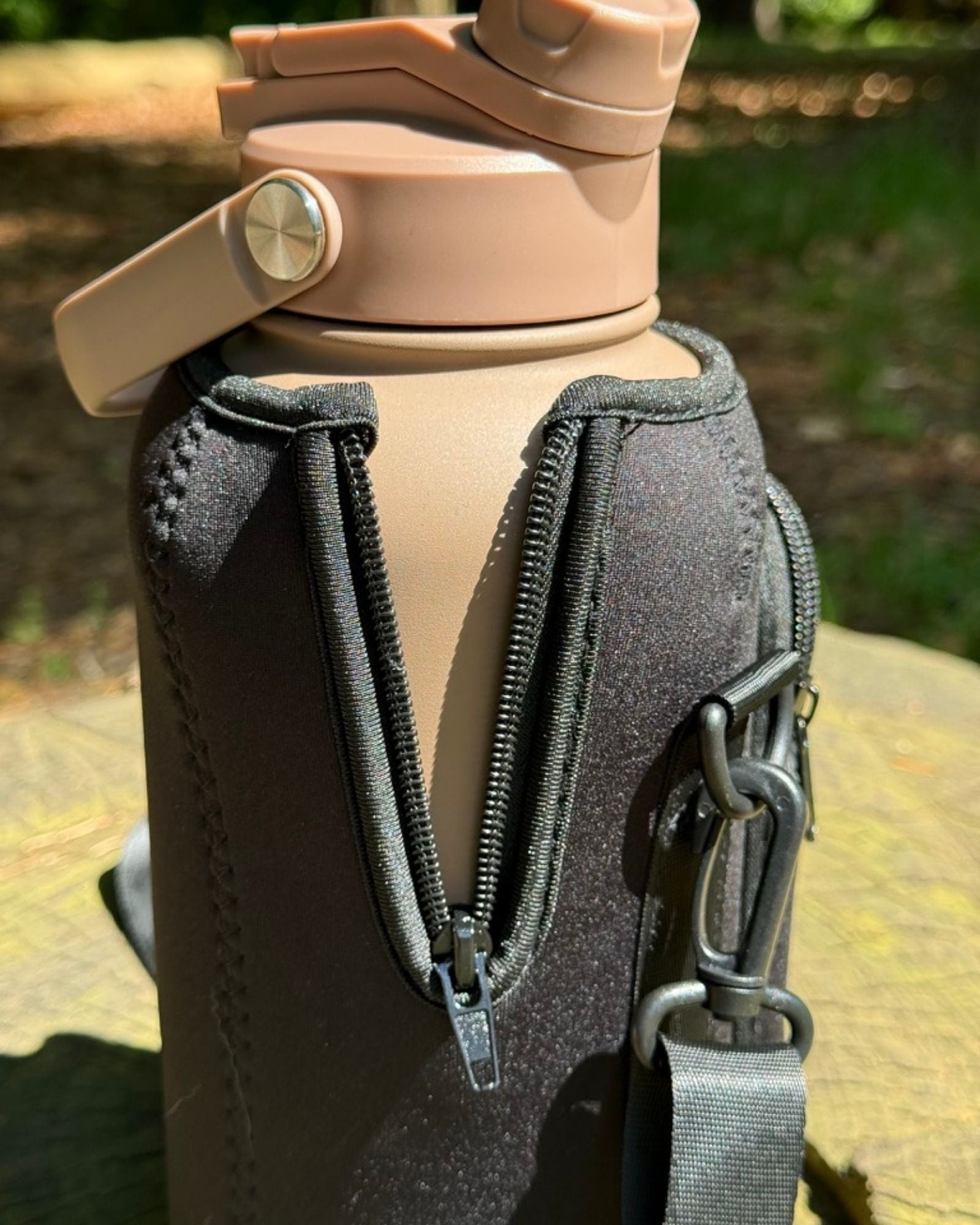 Close-up detail of the black OMNISE water bottle sling showing the durable side zipper stretching to accommodate a tan RoamWell bottle, highlighting the neoprene material and strap hardware.